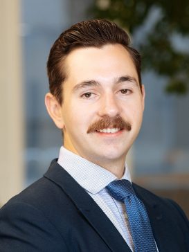 Attorney Jackson Morawski wears a suit and blue tie. He is smiling and has a mustache while standing inside near bright windows and a false tree.