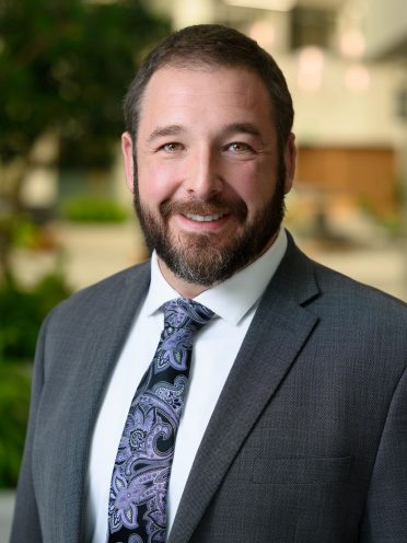 Jed Ullirch is wearing a gray jacket and his tie has a light purple floral design. He has dark brown hair and a short beard.