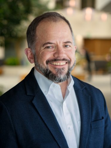 Alex is wearing a white button up shirt with a navy jacket. He has a salt and pepper beard and a low haircut.