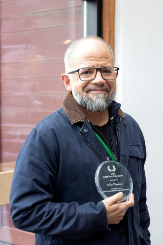 Alex Cleghorn stands in front of a brick wall while holding a clear, circular award for the Judge Nora Guinn Award. He is wearing a dark blue jacket with brown collar, black frame glasses, and has a gray beard.