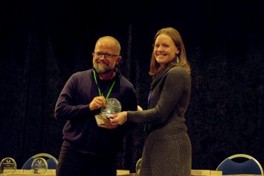 Alex Cleghorn and Danielle Bailey stand on a stage and together hold a clear, circular award for the Judge Nora Guinn Award.