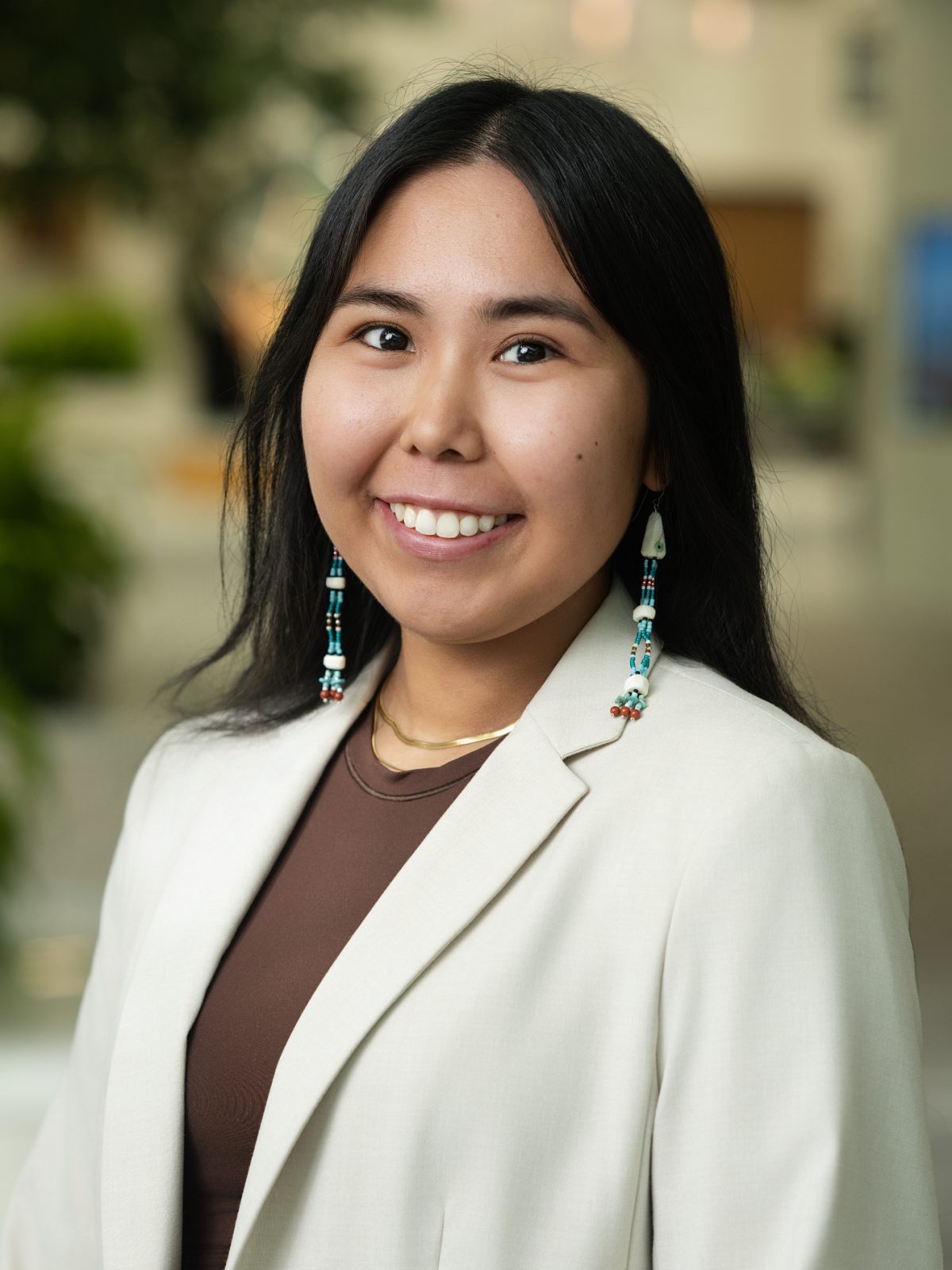 Chanel wears a cream blazer over a brown blouse. She has long black hair and is wearing Alaska Native styled dangly earrings.