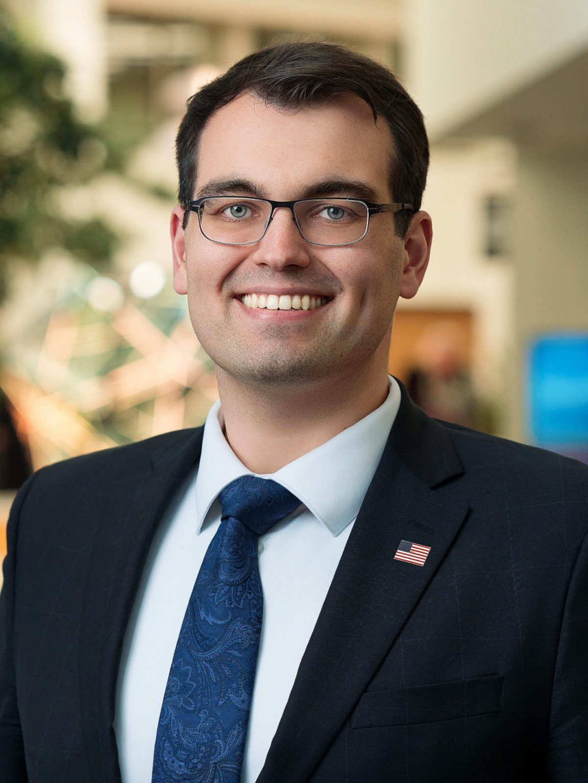 Henry is Smiling and Wearing a Dark Suit and Blue Paisley Tie Wearing Glasses with a Small American Flag Pin on the Lapel