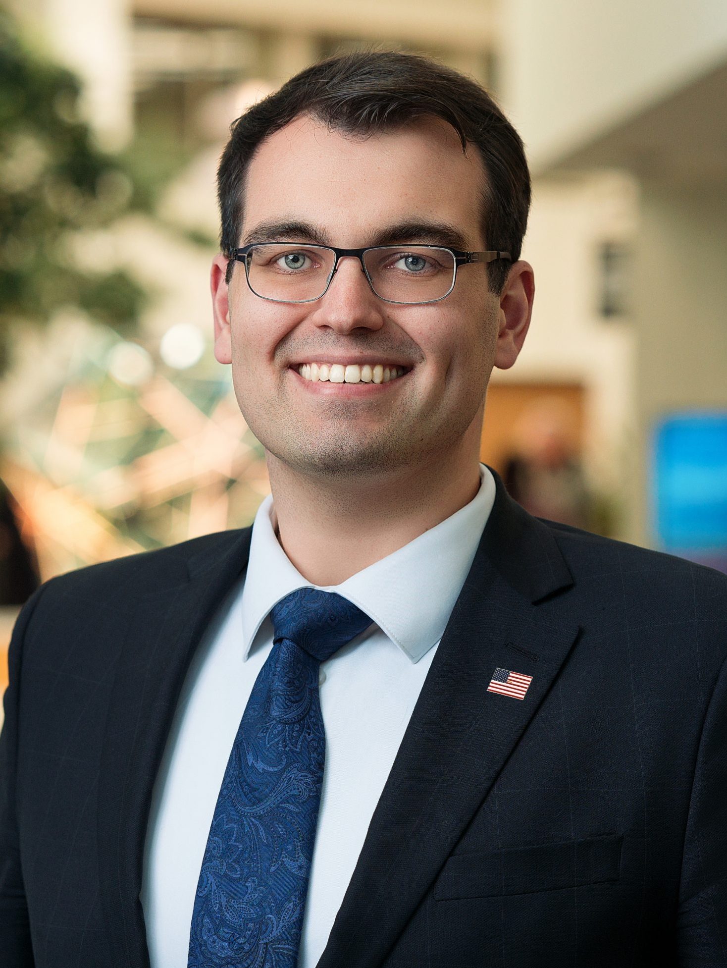Henry is Smiling and Wearing a Dark Suit and Blue Paisley Tie Wearing Glasses with a Small American Flag Pin on the Lapel